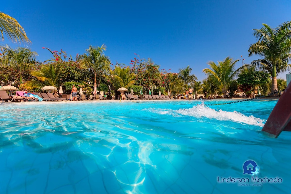 A large outdoor pool is visible, surrounded by palm trees and lounge chairs. The clear blue water reflects the sunny sky, while gentle waves ripple across the surface. Shade is provided by umbrellas placed strategically around the pool area.