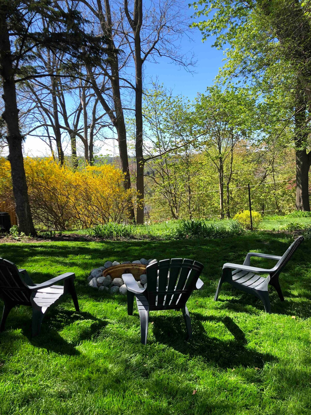 A serene backyard is featured, with a stone fire pit surrounded by three black Adirondack chairs. Lush green grass covers the ground, while trees and bright yellow foliage provide a natural backdrop under a clear blue sky.