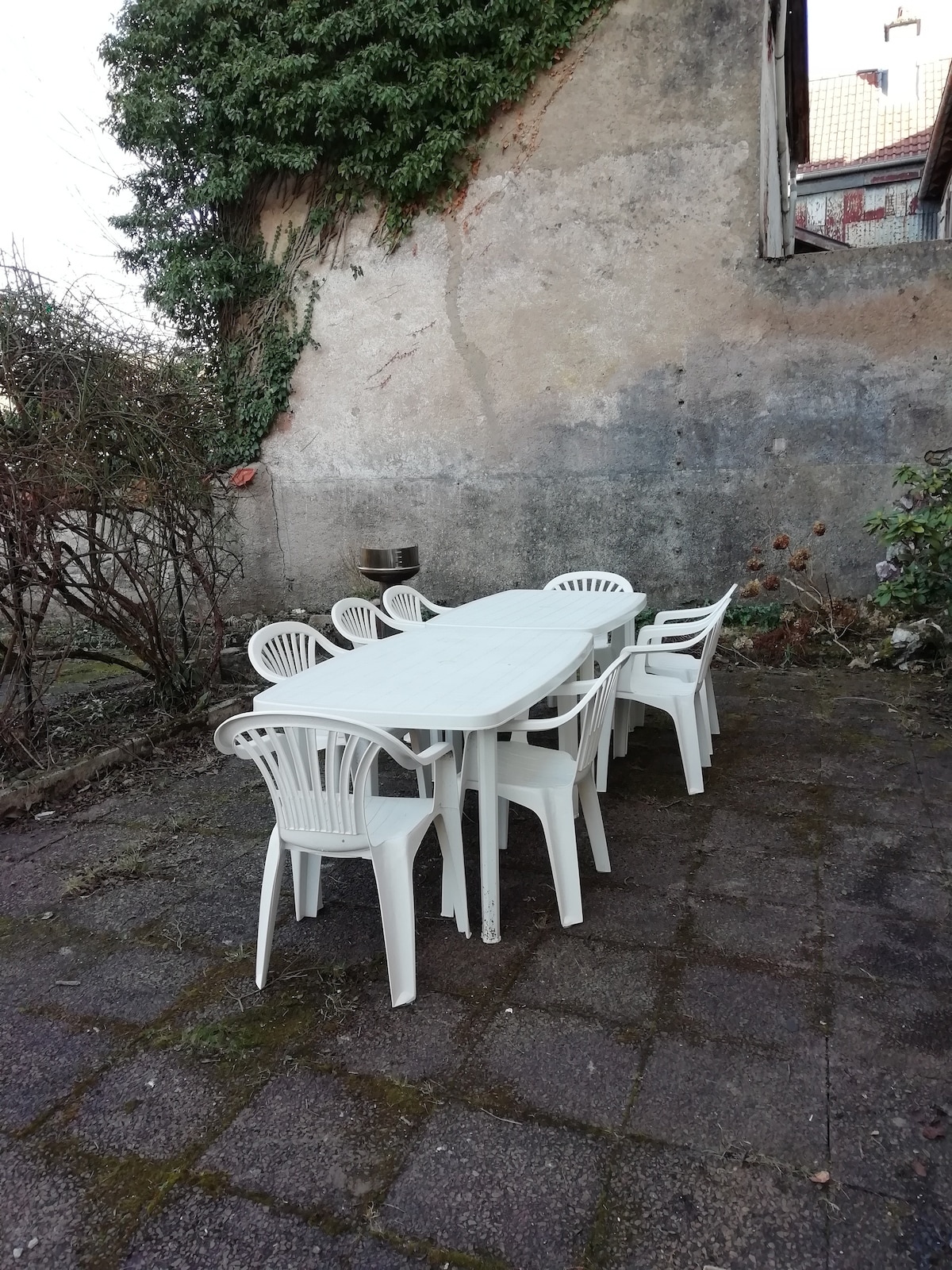 An outdoor dining area is shown, featuring a long rectangular table surrounded by several white plastic chairs. The setting includes a wall partially covered with greenery and a stone surface beneath, creating a simple and functional space for gatherings.