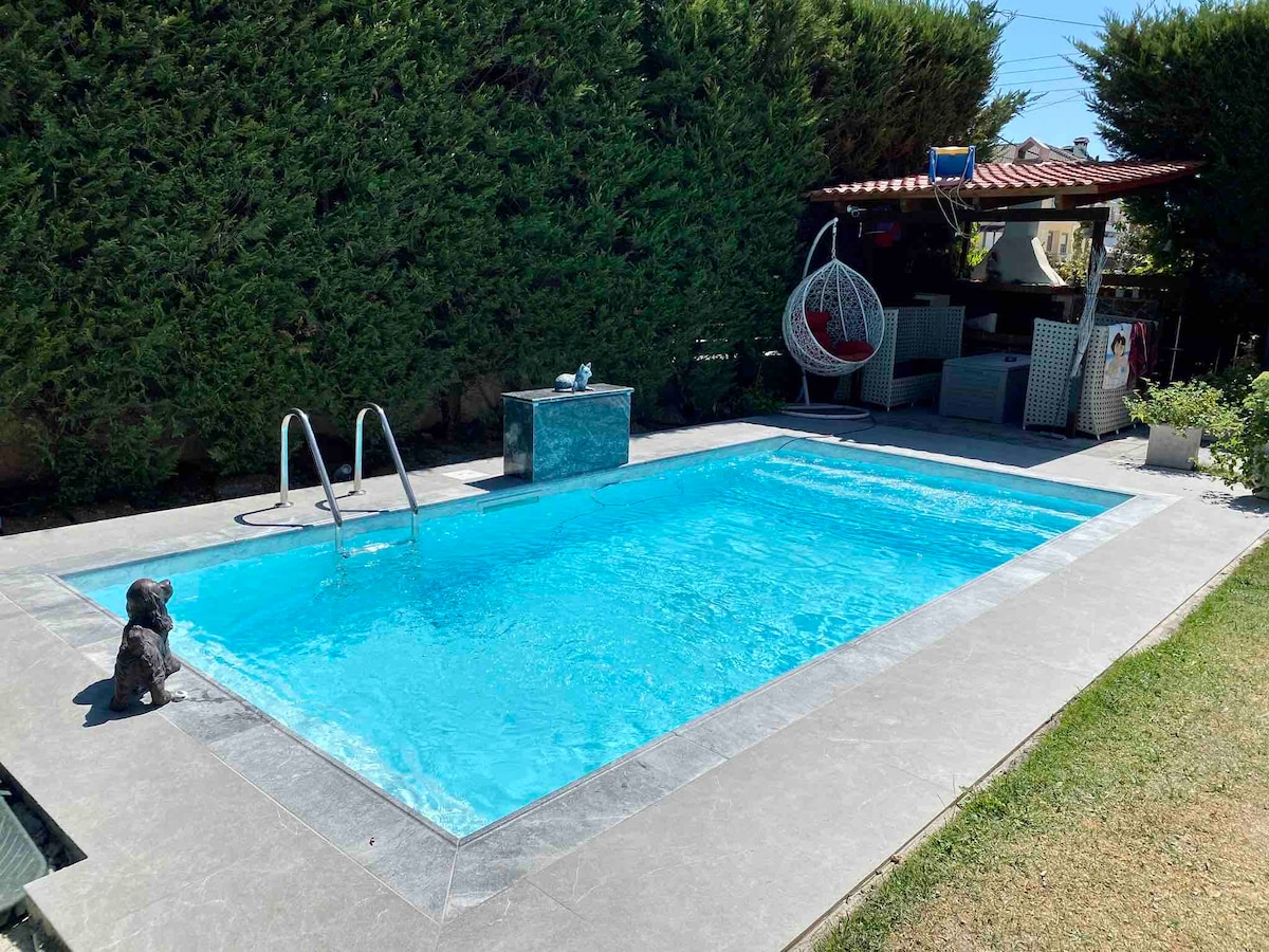 A refreshing outdoor pool is framed by neatly landscaped hedges and smooth gray tiles. Lounge chairs are positioned beside the water, while a covered area features a hanging chair and seating. The serene environment offers a perfect space for relaxation.