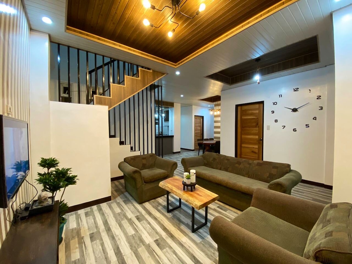 An open living area features two green sofas arranged around a wooden coffee table. Large windows allow natural light to fill the space, while a prominent wall clock adds to the decor. Stairs lead to an upper level, enhancing the room's vertical dimension.