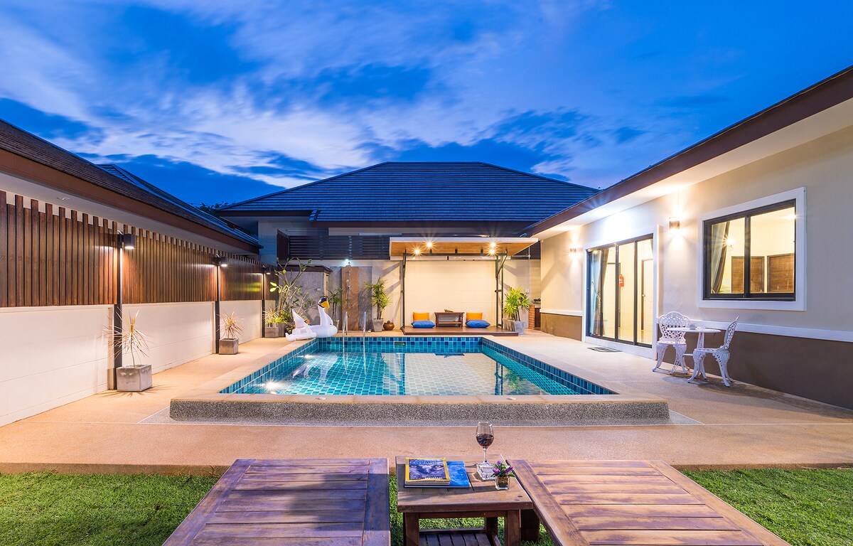 An outdoor area features a private pool surrounded by a sun deck, with seating for relaxation. Evening lights illuminate the space, showcasing the villa's modern architecture. Lush greenery provides a serene backdrop, enhancing the peaceful atmosphere of the surroundings.