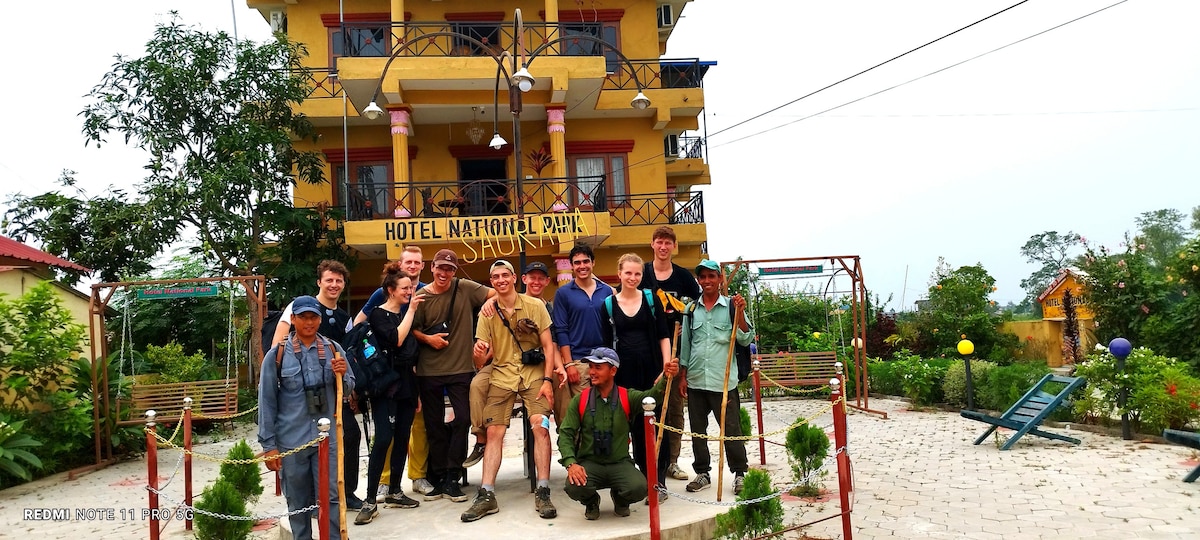 The front of Hotel National Park Sauraha is displayed, featuring a three-story building with a colorful balcony. Guests are gathered on a path lined with decorative plants and railings, with green trees and garden space visible in the background.