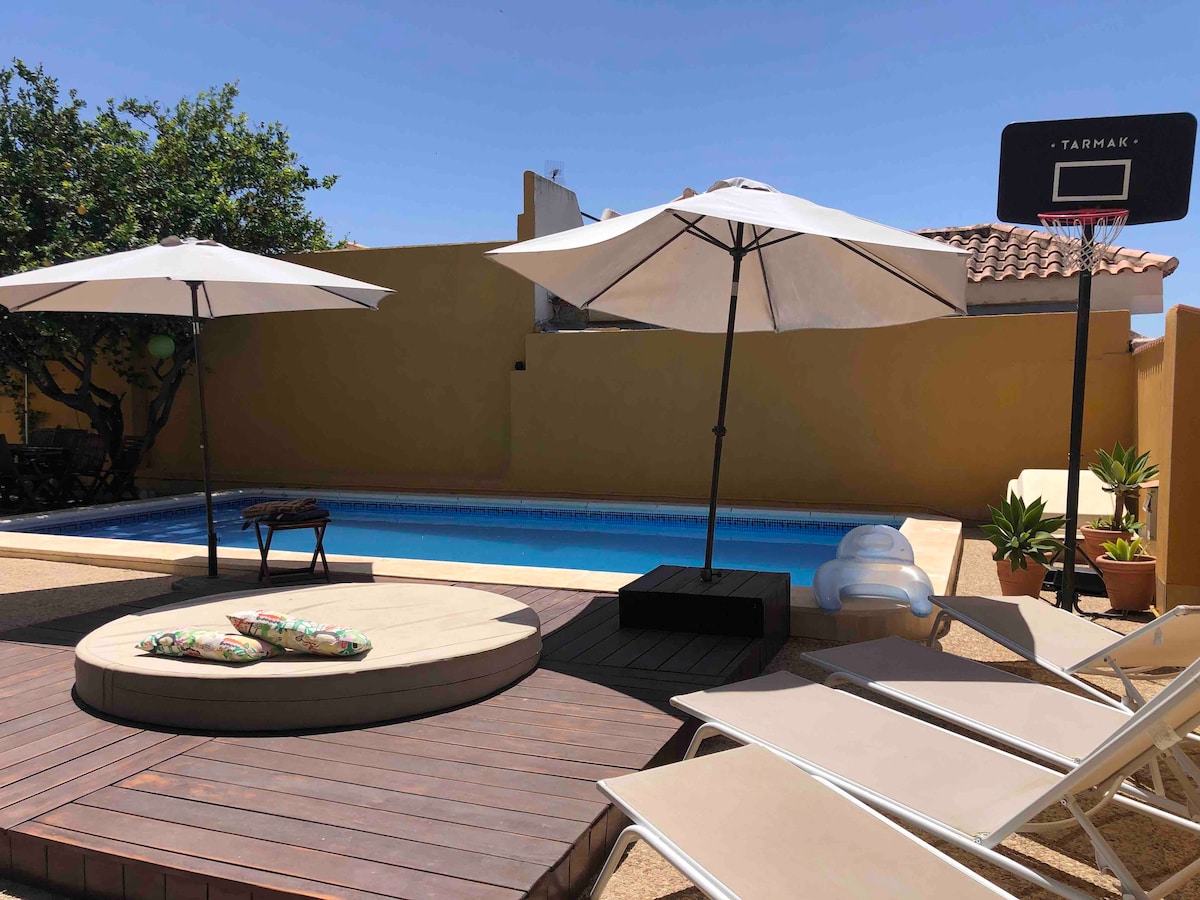 An outdoor area features a private pool surrounded by a wooden deck with lounge chairs. Two umbrellas provide shade over a circular platform. A basketball hoop stands nearby, alongside potted plants that enhance the serene setting.