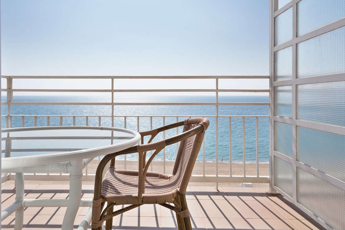 A terrace with a clear view of the sea features a round glass table surrounded by a wicker chair. The calm water reflects sunlight, creating a serene atmosphere. The deck is lined with a railing, enhancing the open and airy feel of the space.