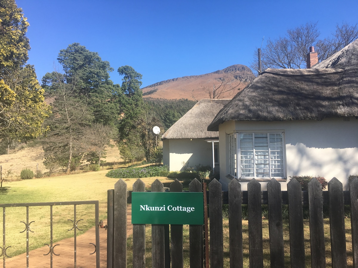 Nkunzi Cottage is viewed from the entrance, featuring a thatched roof and a welcoming sign. The surrounding landscape includes a well-kept lawn and trees, with the backdrop of Inhlosane Mountain showcasing its distinctive peaks under a clear blue sky.