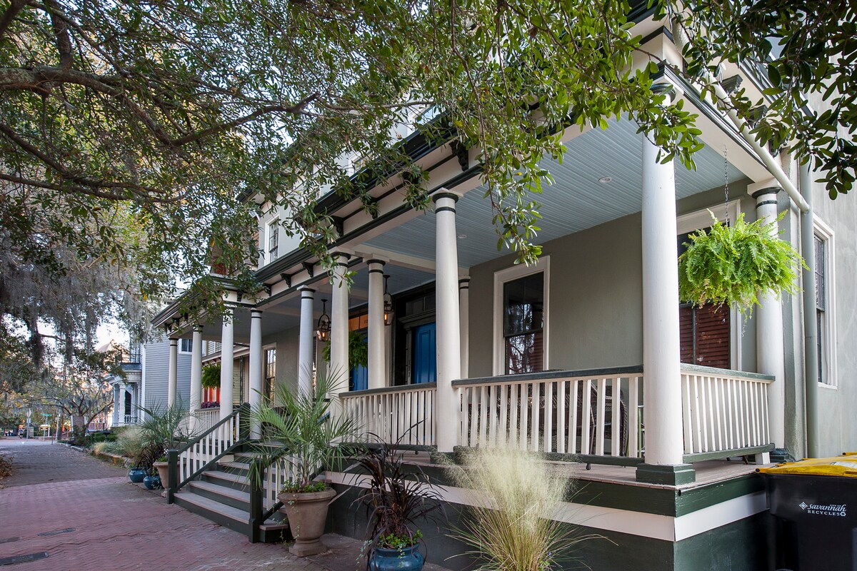 The front exterior of the townhouse displays elegant columns supporting a spacious porch. Lush greenery and potted plants provide a vibrant touch, while the shaded area invites relaxation. Steps lead up to the entrance, framed by natural foliage creating a welcoming atmosphere.