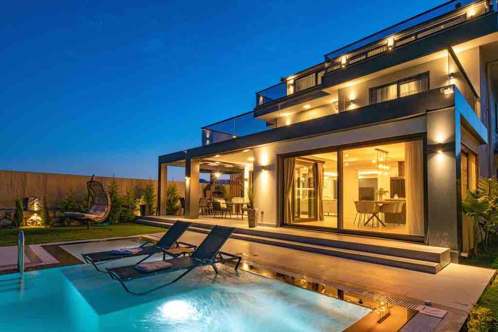 The villa is illuminated at dusk, showcasing a private swimming pool with lounge chairs positioned nearby. Expansive glass doors offer a view into the elegantly designed interior, where a dining area is visible. The outdoor area is surrounded by greenery, enhancing the tranquil setting.