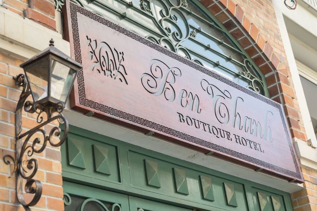 The hotel exterior features a charming sign displaying the name 'Ben Thanh Boutique Hotel', crafted with intricate lettering. A decorative lantern is attached beside the sign, contributing to the building's inviting presentation. The brick facade and ornate green door add character to the entrance.