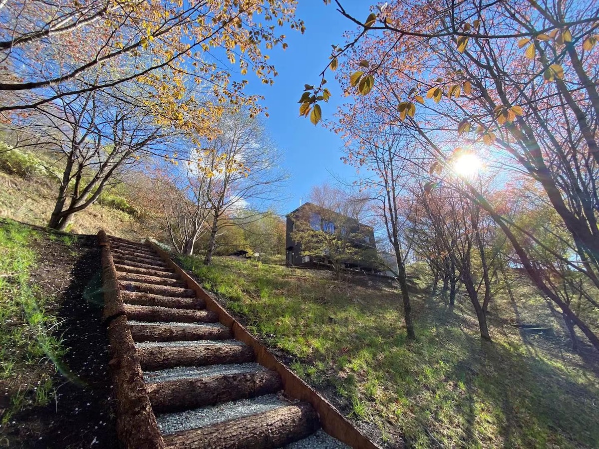 A flight of log steps leads through a lush green landscape adorned with blooming cherry trees, guiding the way to a modern two-story villa nestled among the woods. Sunlight filters through the branches, casting a warm glow over the serene setting.
