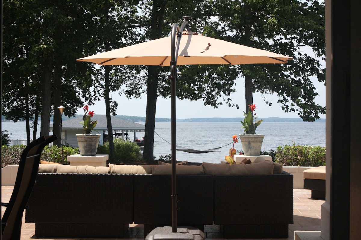 A shaded outdoor seating area features a large umbrella over a comfortable sectional sofa. Potted plants with vibrant flowers are placed nearby, overlooking the serene lake. A hammock can be seen in the distance, inviting relaxation with scenic water views.