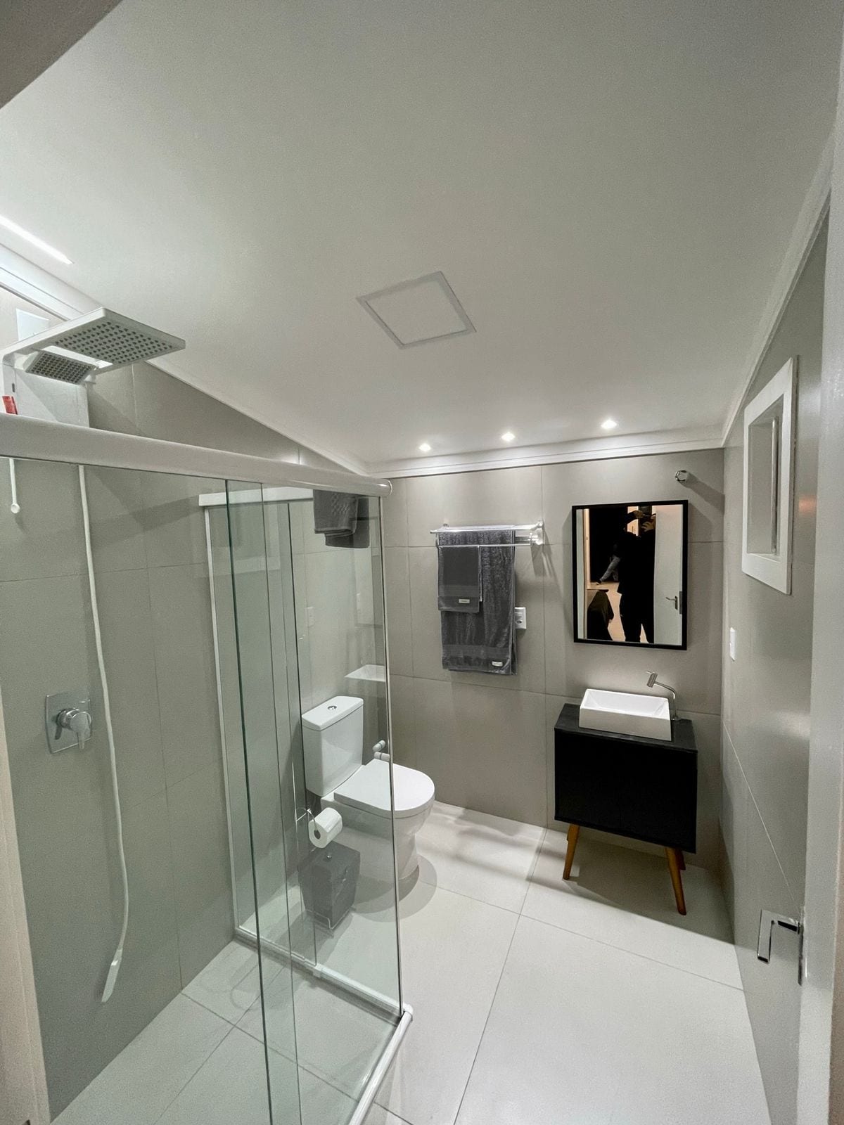 A modern bathroom features a walk-in shower with a glass enclosure. A sleek vanity with a dark cabinet and a minimalist sink is visible. Gray tiles adorn the walls, and a large mirror enhances the space. Soft lighting creates an inviting atmosphere.