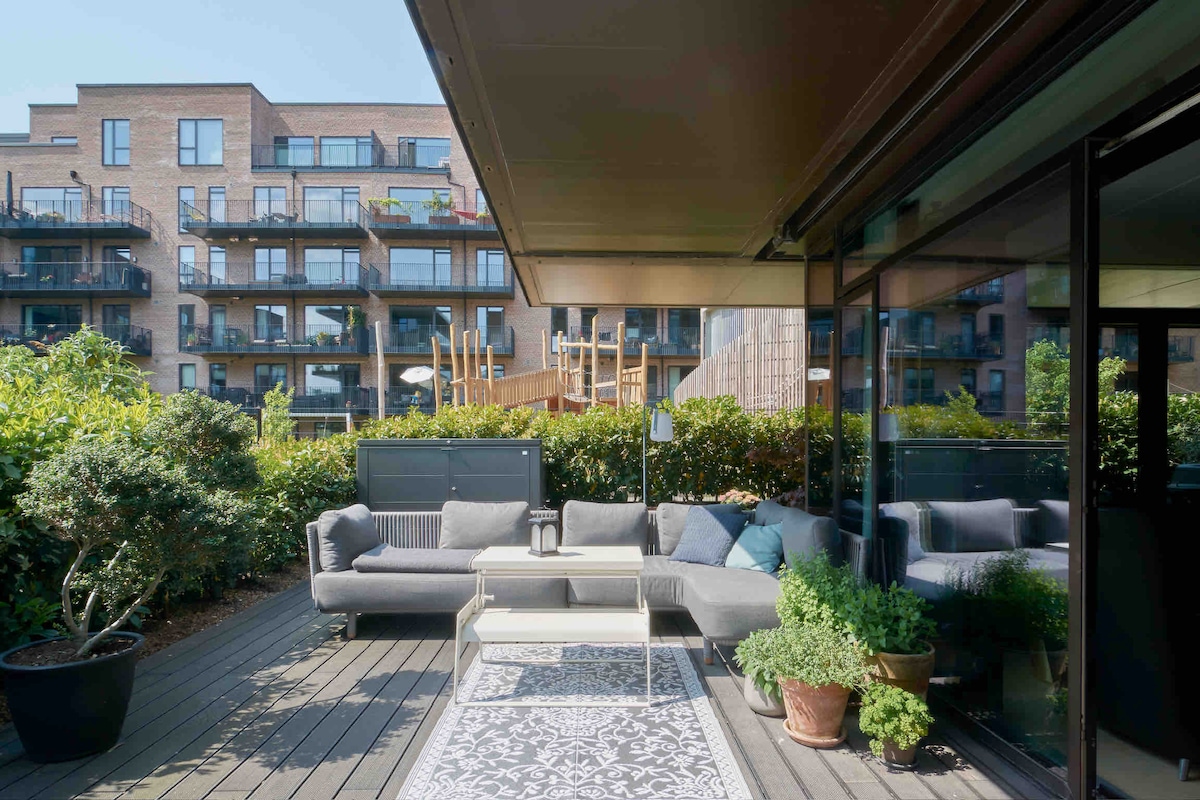 A spacious outdoor terrace is furnished with a comfortable seating area, featuring a mix of grey sofas arranged around a white coffee table. Lush greenery surrounds the space, providing privacy. In the background, a playground is visible, offering a vibrant atmosphere.