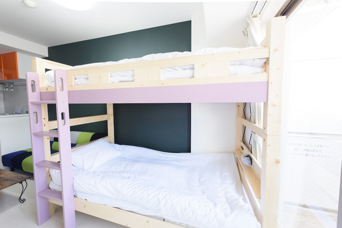 A bunk bed with wooden frames in light tones is showcased against a dark green accent wall. The upper bed is unmade, while the lower bed is arranged with fresh, white bedding. A compact ladder provides access to the top bunk.