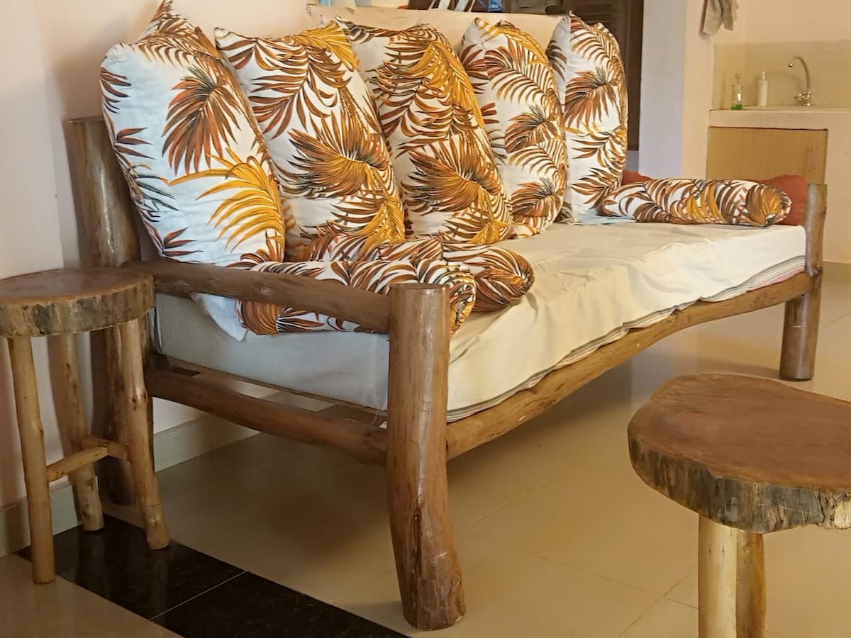 A wooden sofa features a natural finish with plush cushions adorned in a tropical leaf pattern. Two wooden side tables accompany the seating, showcasing a rustic design. The soft color palette blends harmoniously with the light surroundings.