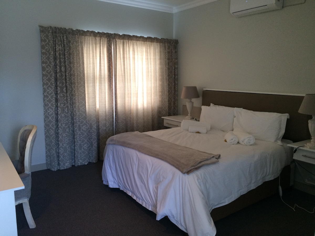 A spacious room features a large bed with soft linens, complemented by a warm-toned throw. Two bedside tables with lamps flank the bed, and a writing desk is positioned nearby. Natural light filters through elegant curtains, enhancing the serene ambiance.