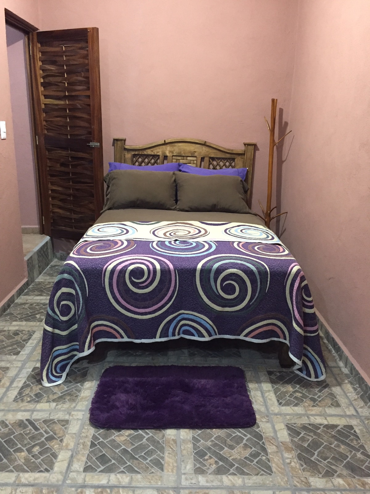A compact bedroom is shown with a bed covered in a patterned blanket featuring swirling designs. Two dark pillows rest against the headboard. A plush rug in a deep purple tone is positioned on the floor, and a wooden coat rack stands in the corner.