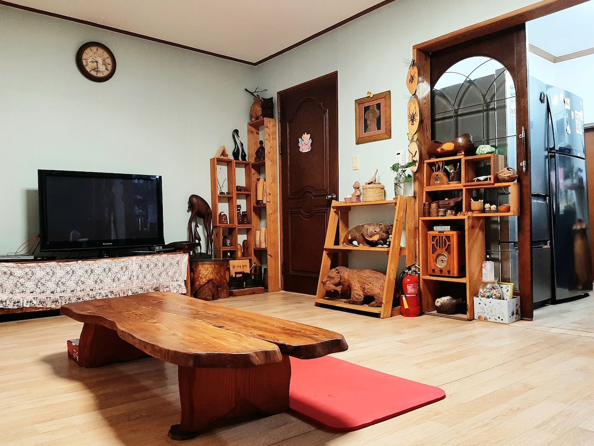 The living area features a wooden low table positioned on a red mat, surrounded by a variety of decorative wooden sculptures on shelves. A large television is mounted on the wall, and a wooden cabinet displays additional art pieces. Natural light fills the room from a nearby window.