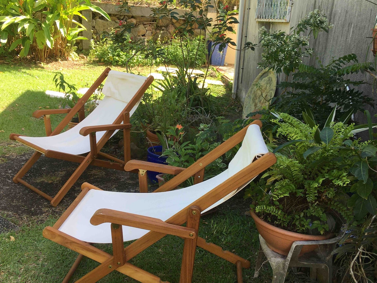 Two wooden lounge chairs are positioned on a grassy area within a lush garden. Surrounding foliage includes various plants and flowers, enhancing the serene outdoor environment. A terracotta pot with greenery adds to the tranquil setting, providing a relaxing space for guests.