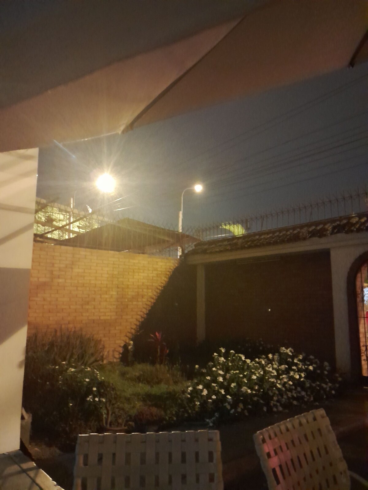 An outdoor area is depicted in low light, showcasing a lush garden with white flowers. A brick wall serves as a backdrop, and the scene is complemented by streetlights illuminating the surroundings. Chairs are positioned nearby, fostering a relaxed atmosphere.