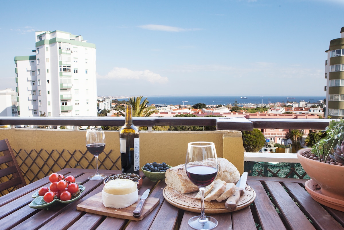 A wooden table is set on the balcony, featuring a selection of tomatoes, cheese, olives, and slices of bread, accompanied by a bottle of wine and glasses. The backdrop includes a clear view of the coastline and nearby buildings, enhancing the relaxed outdoor dining experience.