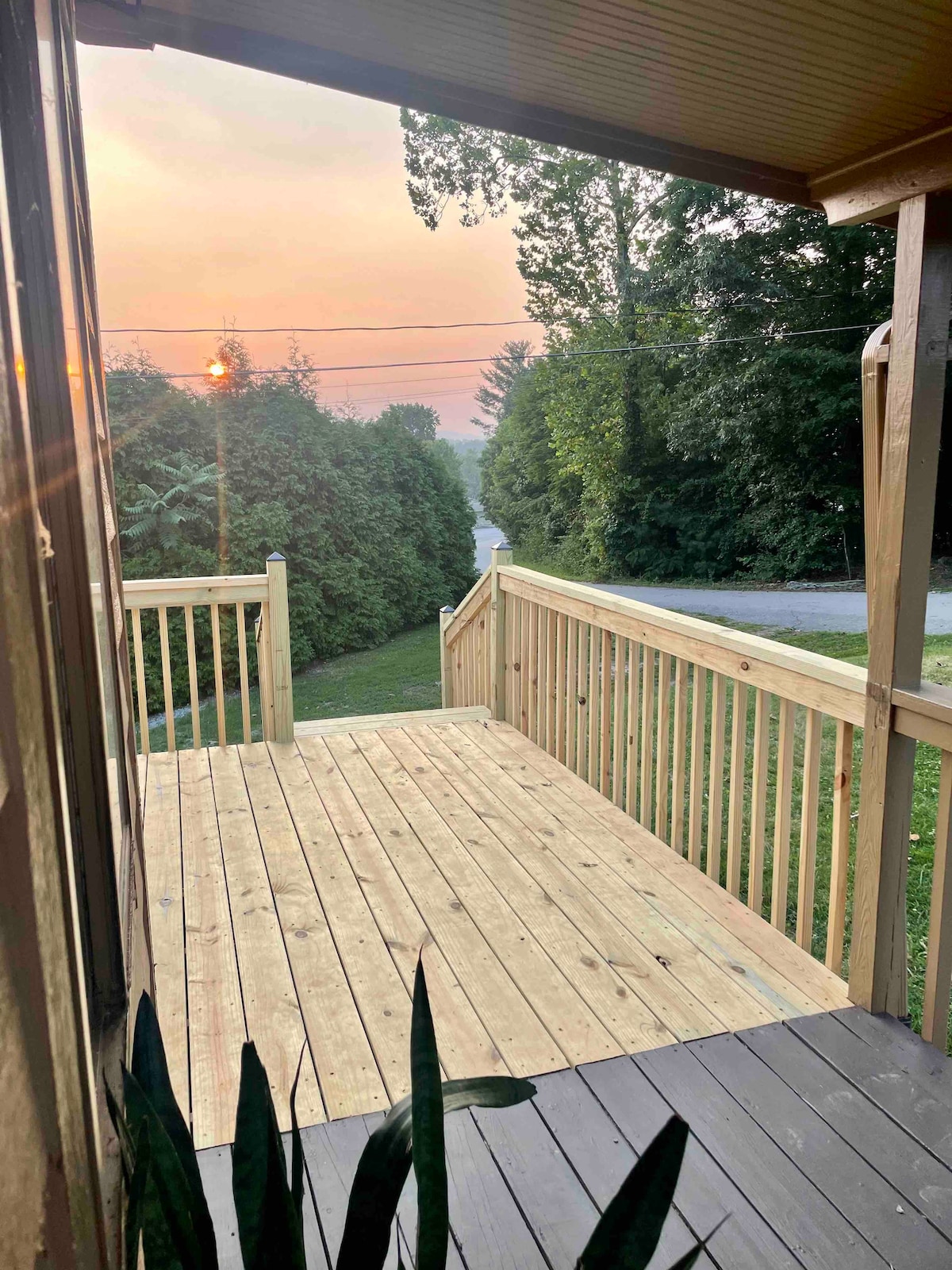A wooden deck is showcased, featuring a smooth, light-colored surface. Green trees line the pathway in the background, with the sun setting beyond them, casting a warm glow over the scene. The railing provides a sense of openness to the surrounding natural area.