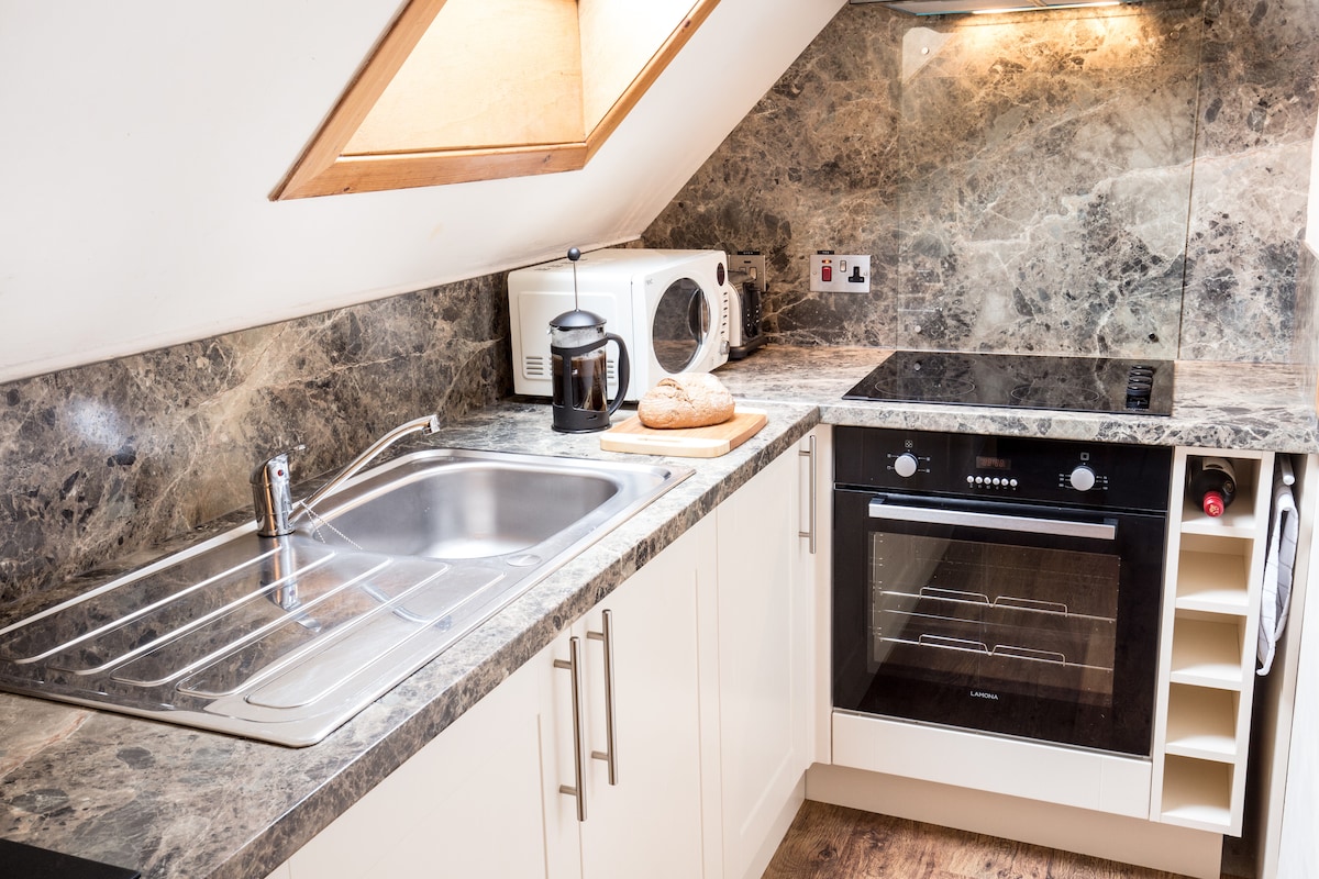 A compact kitchen area is showcased with a marble countertop. An electric oven and stove are situated next to a microwave. A kettle and bread sit on a wooden cutting board. Cabinets and a sink complete the well-equipped space, illuminated by natural light.