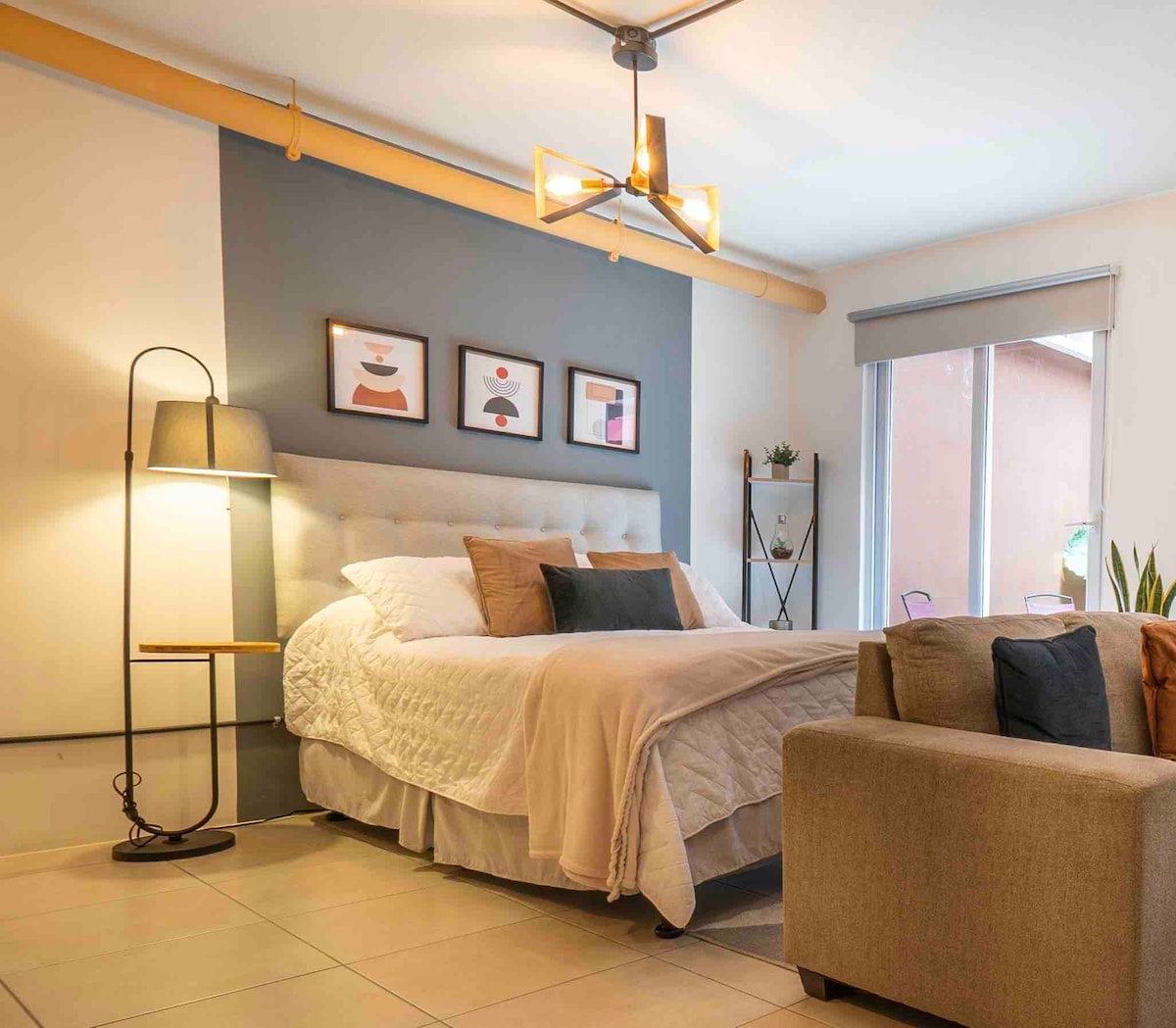 A well-appointed sleeping area features a spacious bed topped with neutral-colored bedding and decorative pillows. Artwork is displayed on the wall above the bed. A lamp stands beside the bed, providing soft illumination, while a small table and plants are visible in the background.