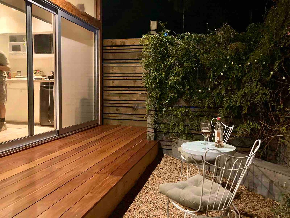 An outdoor seating area features a small round table surrounded by two white wire chairs. A wooden deck is visible, with a lush green wall in the background. Soft lighting is present, contributing to a serene ambiance.