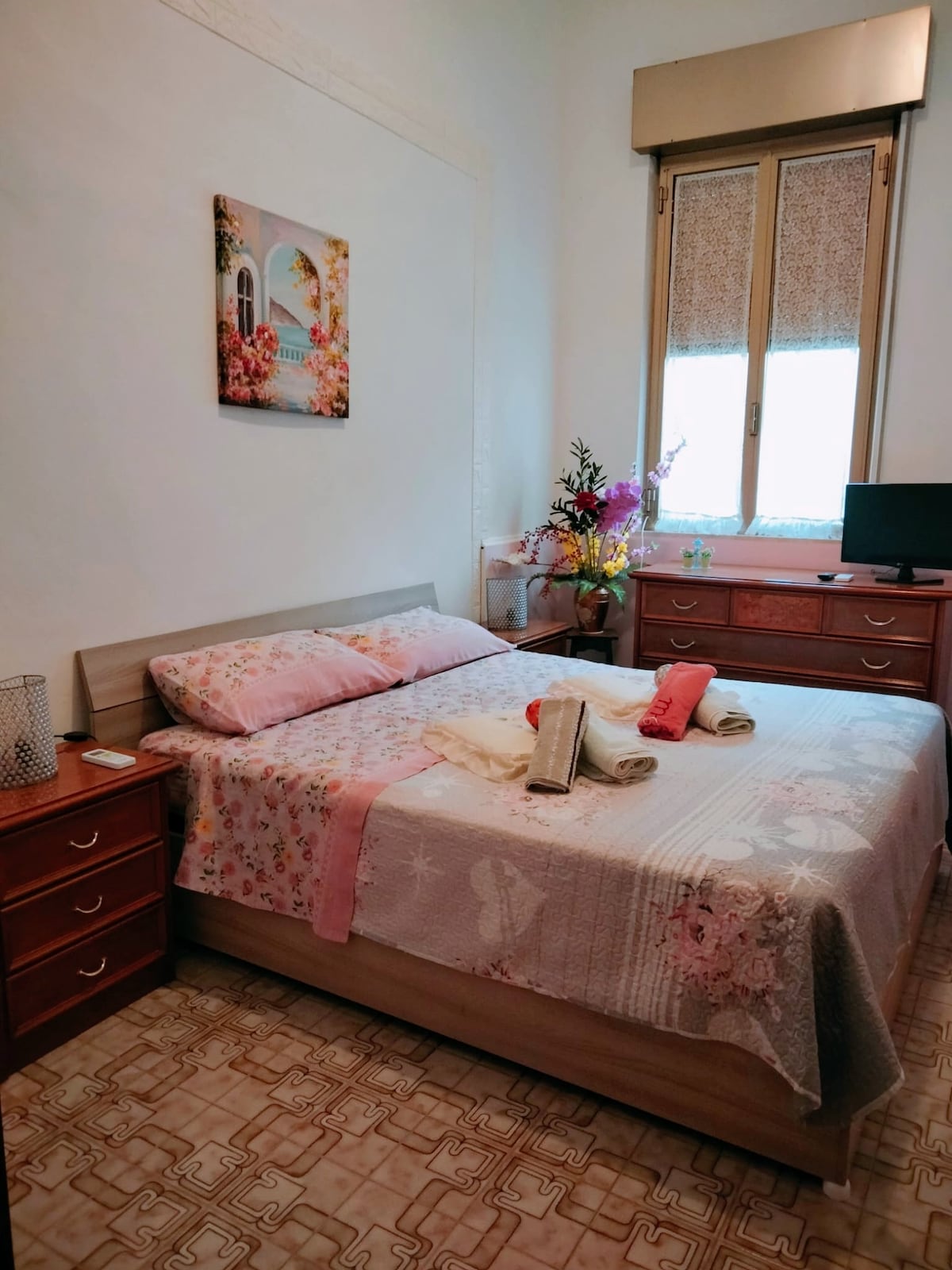 A welcoming bedroom is furnished with a spacious bed, adorned with a floral-patterned coverlet. Natural light filters through a window, illuminating the room. A wooden dresser holds decorative items, and a small television is positioned nearby, enhancing the comfort of the space.