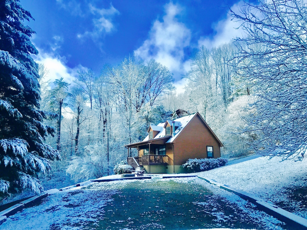 A cozy cabin is set against a backdrop of snow-covered trees and a clear blue sky. A gentle slope leads up to the cabin, which features a wooden porch and multiple windows, inviting natural light during the daylight hours.