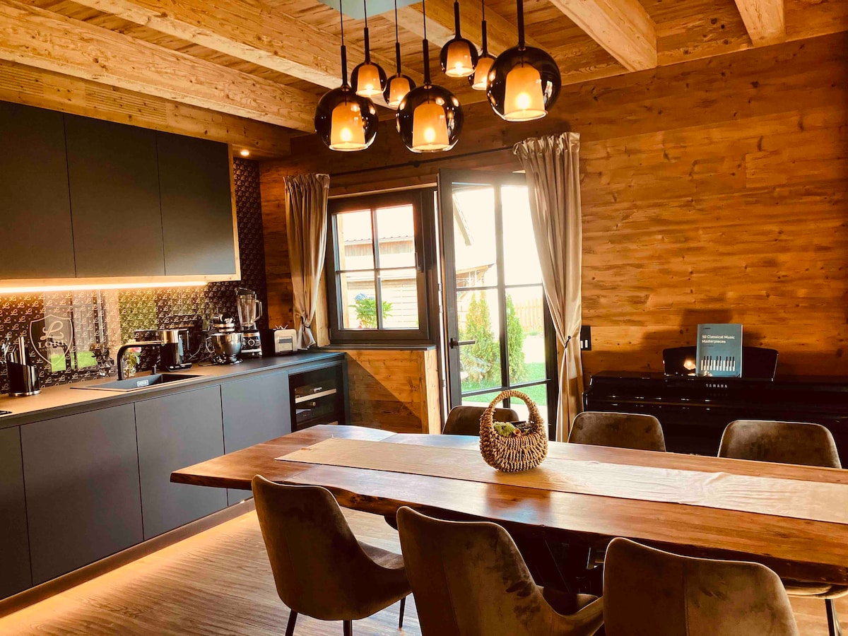 A modern kitchen is paired with a spacious dining area, featuring a large wooden table surrounded by comfortable chairs. Pendant lights illuminate the space, while large windows provide natural light and views of the outdoor greenery. The kitchen is equipped with sleek appliances and stylish cabinetry.