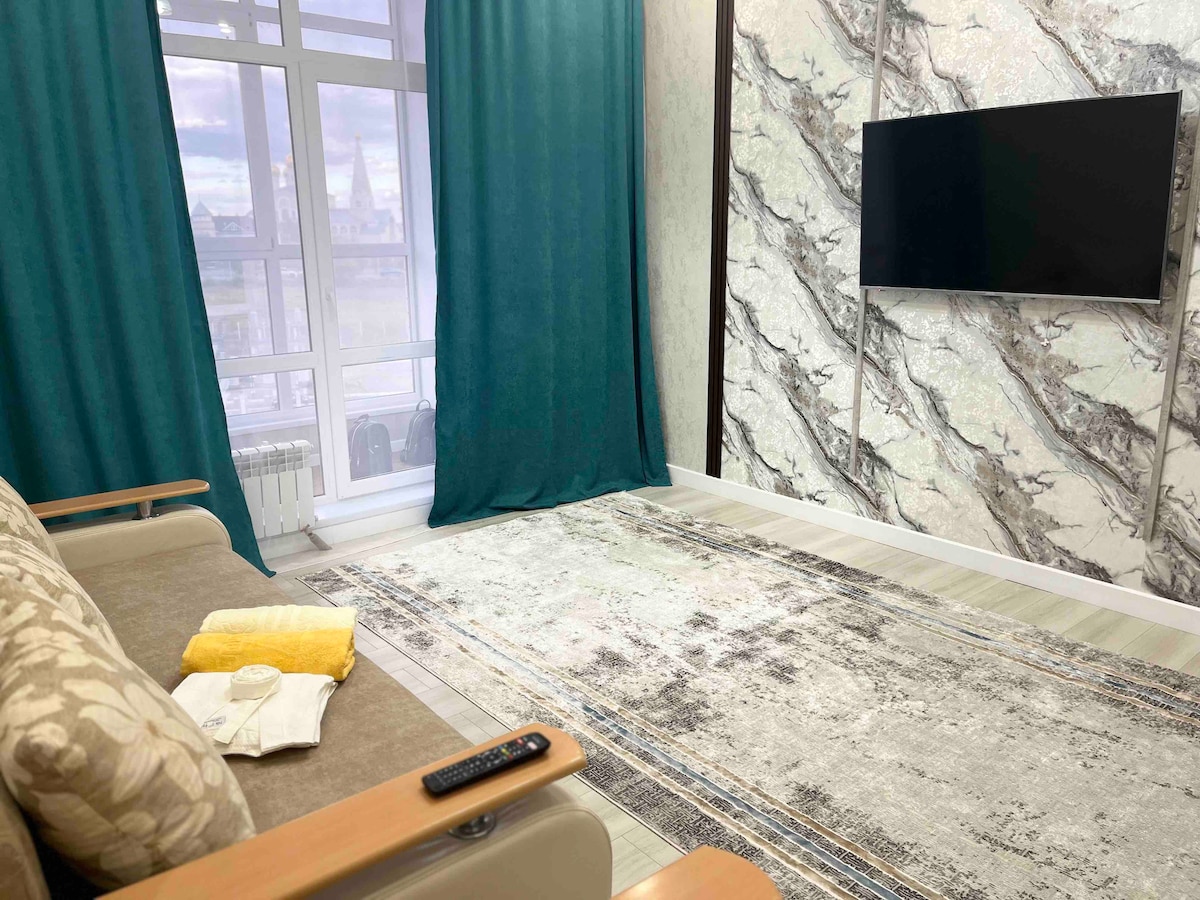 A cozy living area features a comfortable sofa with decorative cushions. A sleek television is mounted on a stylish wall, while large windows dressed with teal curtains allow natural light to fill the space. A patterned rug adds texture to the floor.
