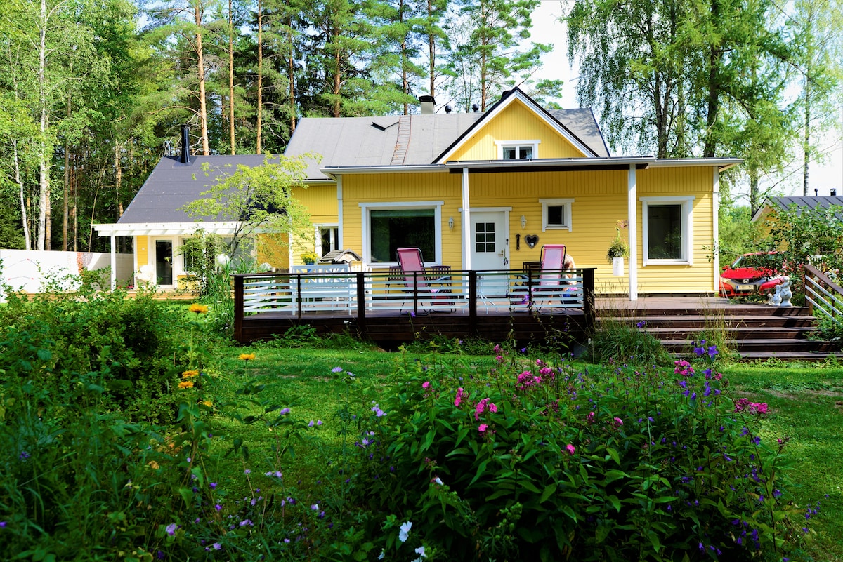 A bright yellow house is set amidst lush greenery, featuring a spacious deck with pink chairs. Surrounding flower beds add color to the scene. The property is bordered by tall trees, offering a peaceful atmosphere.