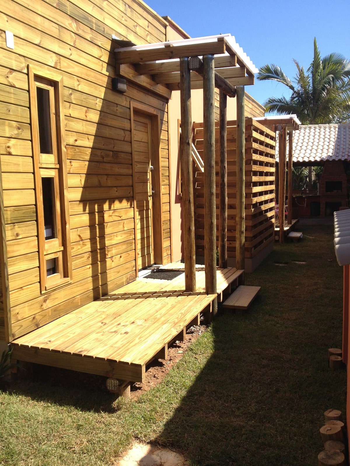 A wooden deck extends toward a rustic building, featuring vertical wooden slats and large windows. Sunlight casts shadows on the grass, highlighting the natural materials and serene environment. Surrounding palm trees provide a tropical ambiance.
