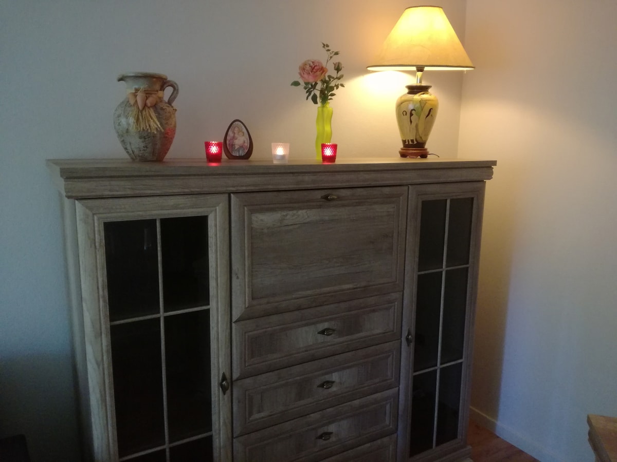 A wooden cabinet with glass doors is featured, showcasing a decorative vase and a small framed picture. A lamp stands on top, casting a gentle light on a small bouquet of flowers. Two lit candles add a touch of warmth to the arrangement.