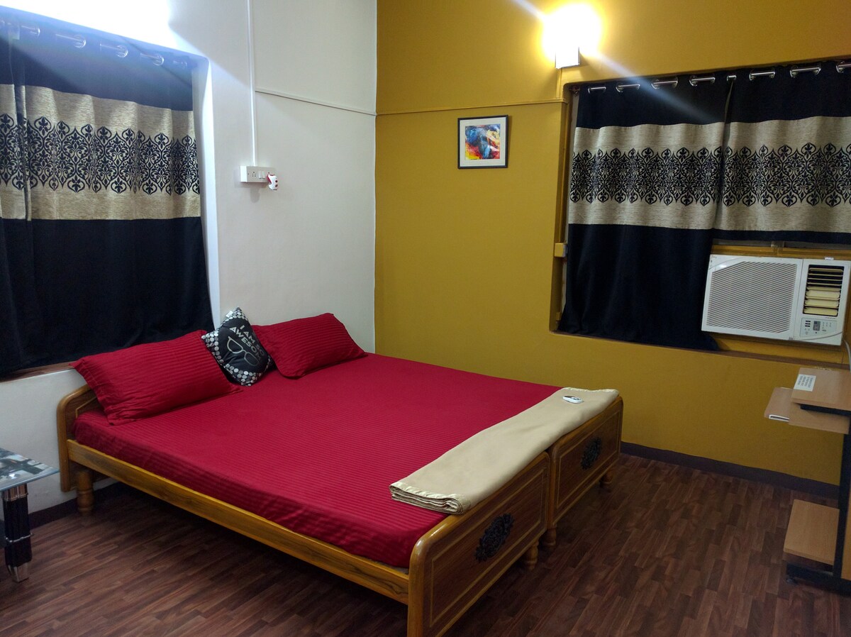 A cozy master bedroom features a wooden bed dressed in red bedding. A bedside table and a wall-mounted air conditioning unit are visible. Dark curtains partially cover the windows, and a wall-mounted artwork adds a personal touch to the space.