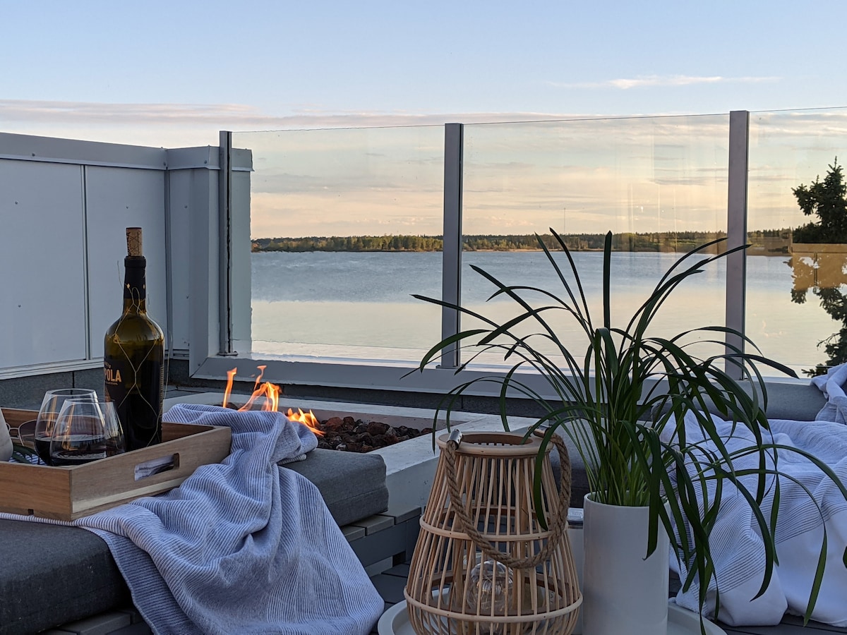A rooftop view showcases tranquil waters and a clear sky. A seating area with soft cushions is complemented by a fire feature. A bottle of wine and glass are arranged on a tray, alongside a decorative lantern and a potted plant, enhancing the inviting setting.