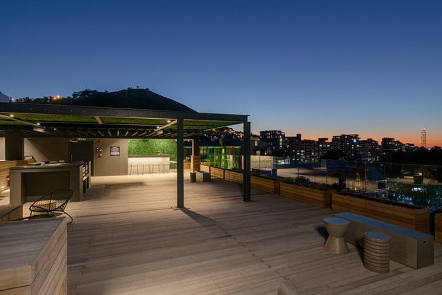 A spacious rooftop terrace is visible, featuring wooden decking and a covered seating area. BBQ facilities can be seen, with the outline of Table Mountain in the background. City lights twinkle in the distance as the sky transitions from dusk to night.