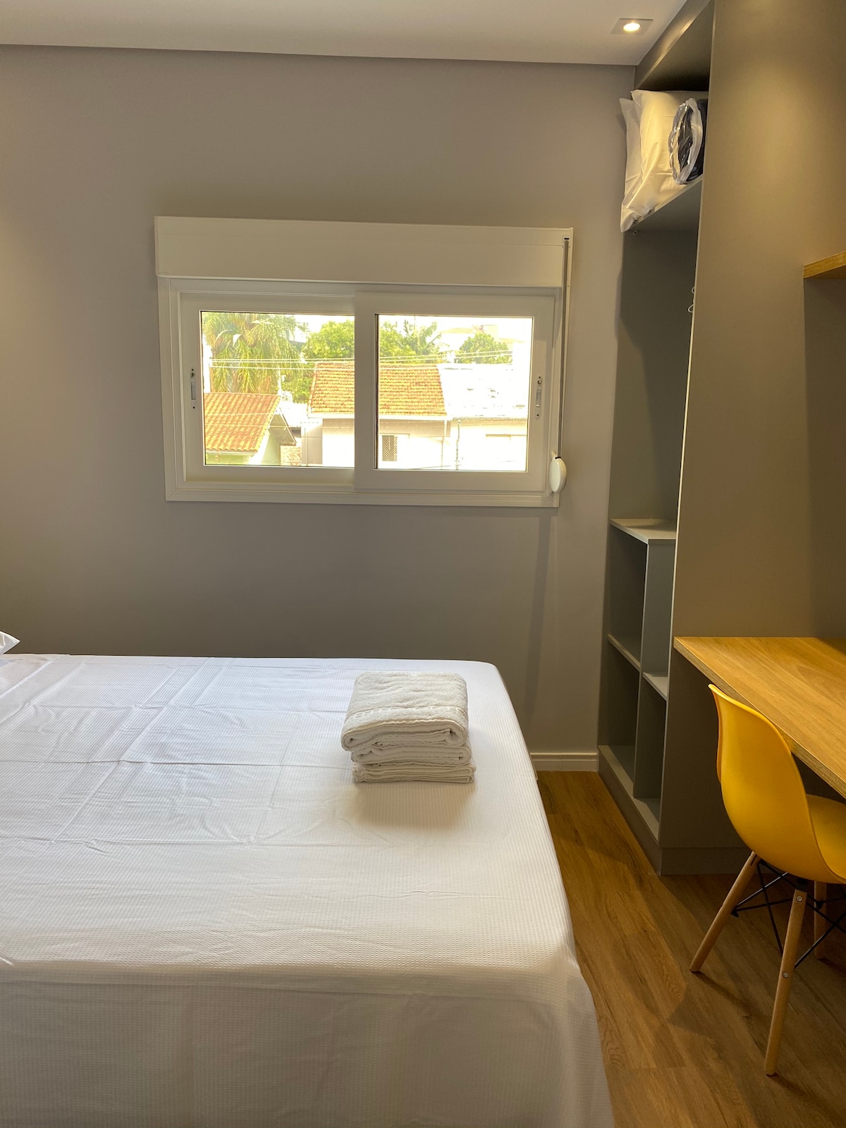 A bedroom features a spacious bed dressed in crisp white linens, with neatly folded towels placed at the foot. Natural light enters through a large window, illuminating a small workspace nearby with a wooden desk and a chair. A closet is positioned along the wall.