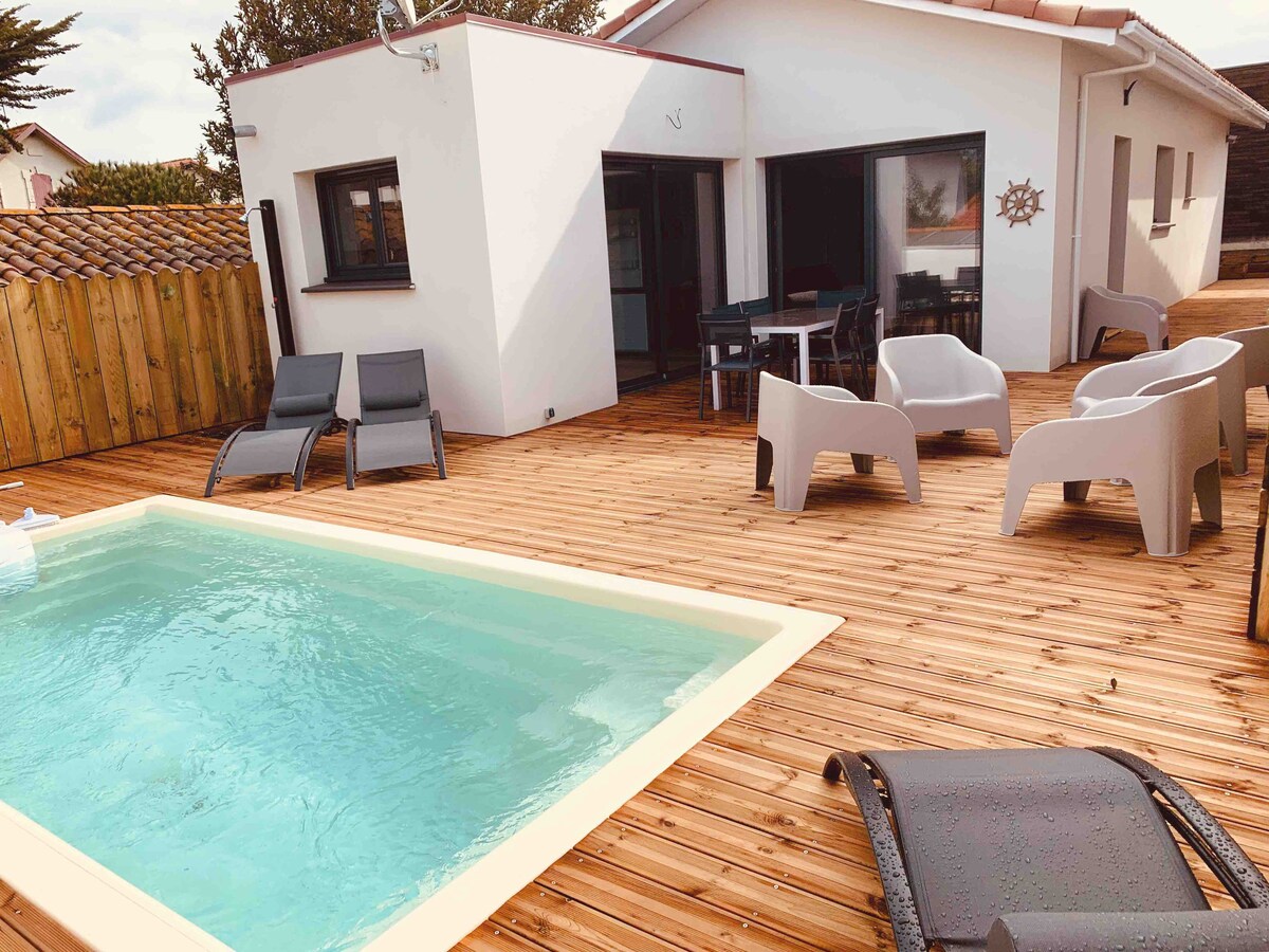An outdoor area is depicted with a small covered pool surrounded by a wooden deck. Several lounge chairs are positioned along the deck, while a dining table with chairs is visible near the house. The exterior walls of the house are finished in white.