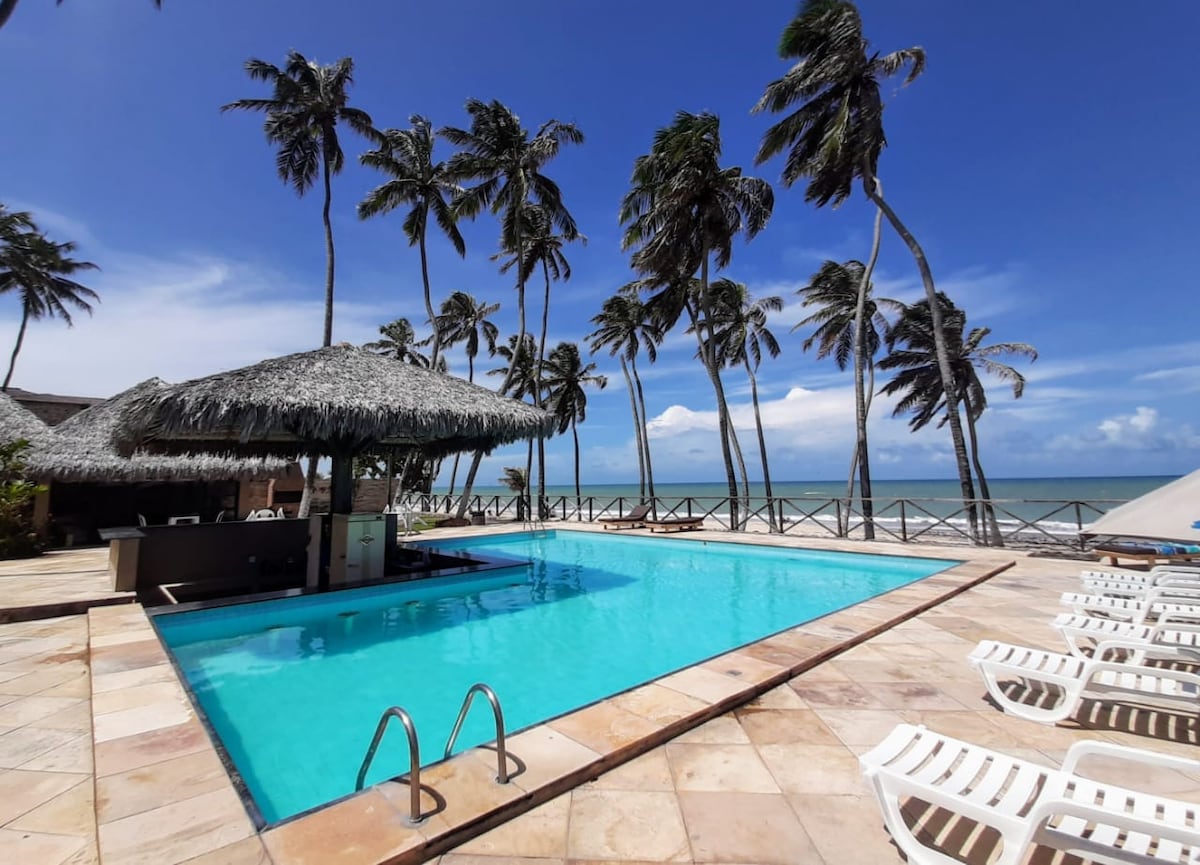 A swimming pool is framed by palm trees, with a thatched roof structure nearby. Lounge chairs are arranged around the pool area, which reflects clear blue skies. The ocean is visible in the background, offering a serene coastal view.