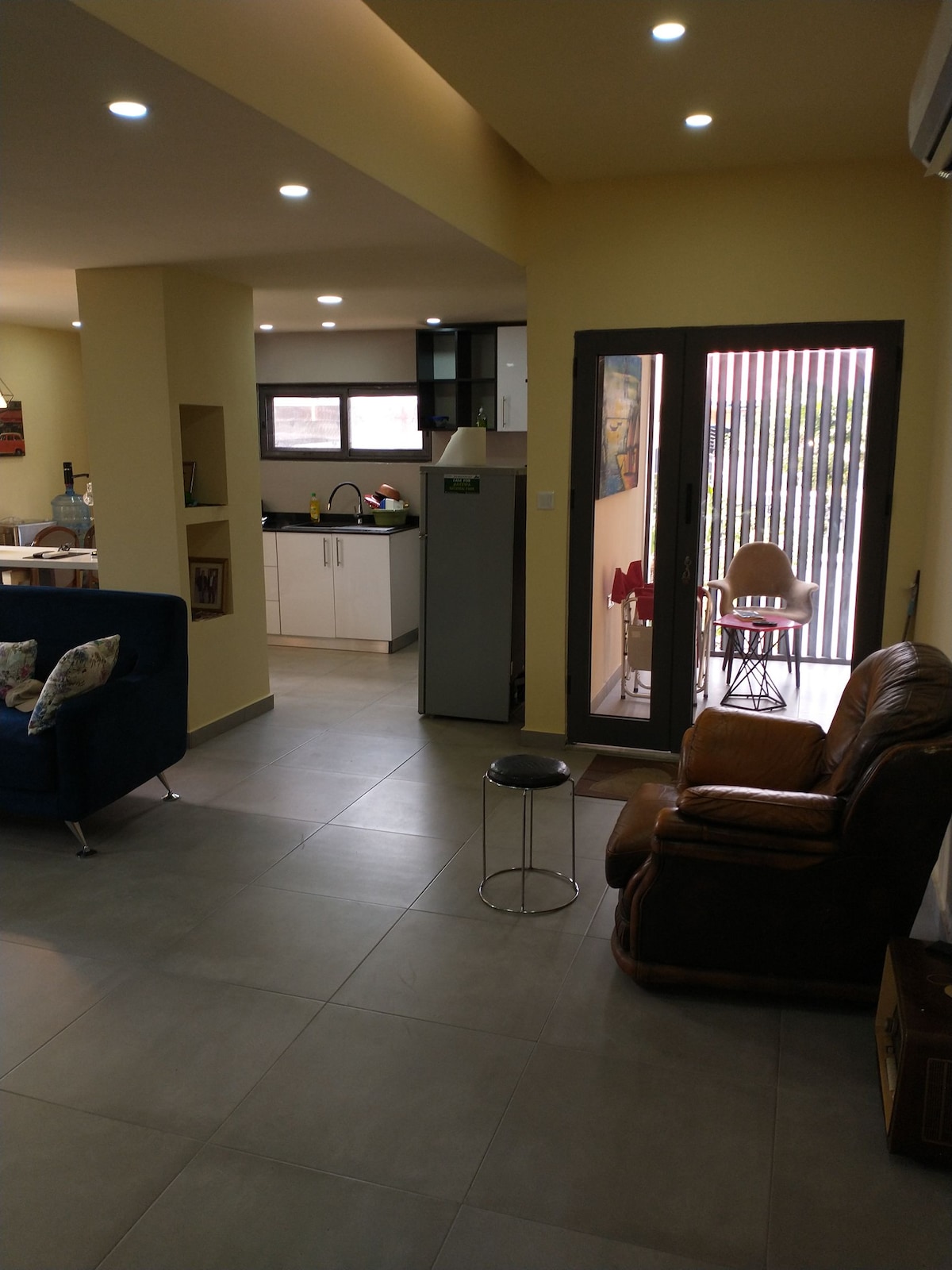 An open layout is featured, showcasing a light-filled living space with a view of the kitchen. A cozy seating area is set near a doorway leading to an outdoor space. Warm yellow walls complement modern light fixtures, enhancing the contemporary ambiance.
