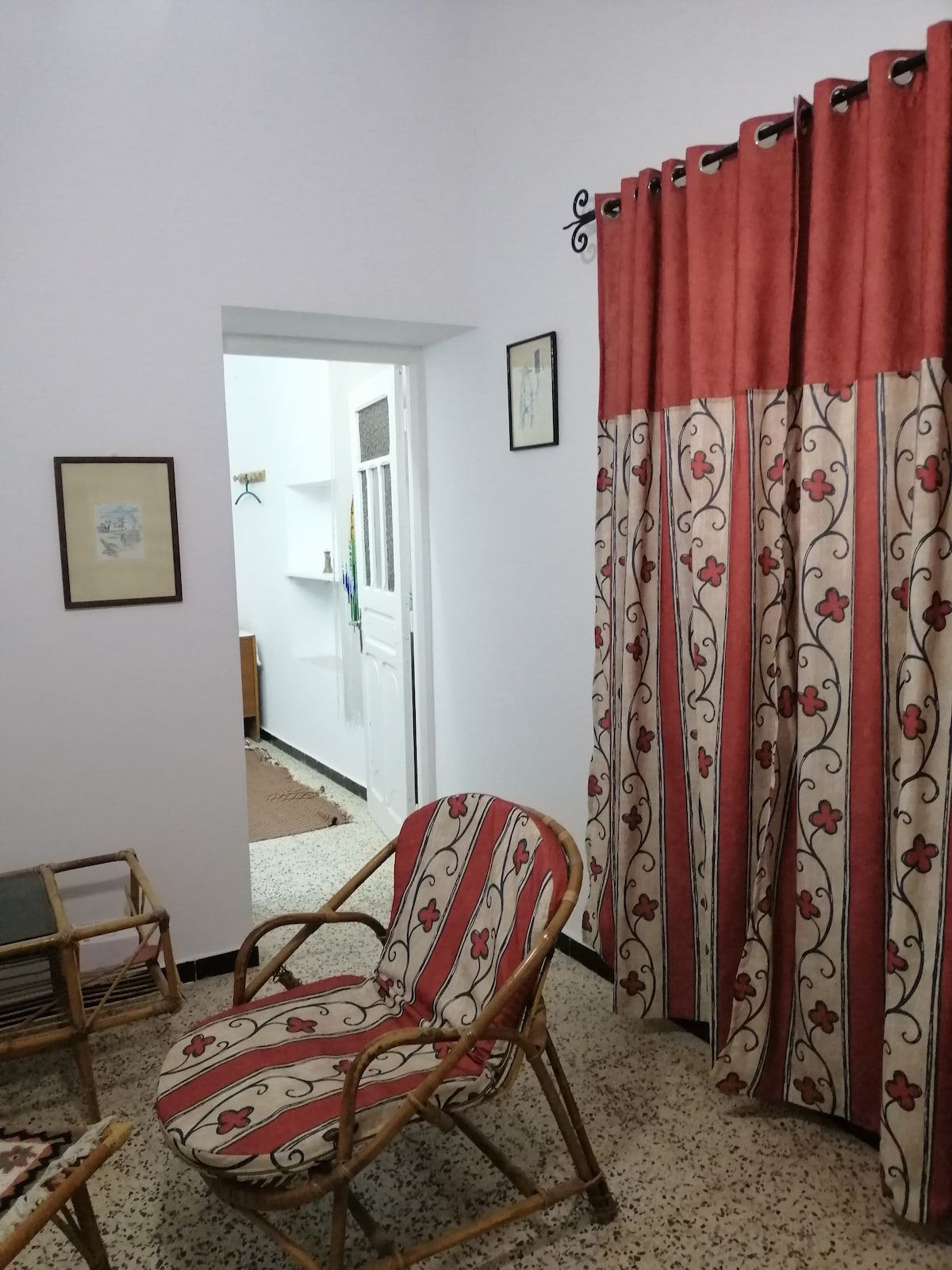 A cozy corner features a rattan chair with a floral-patterned cushioned seat. Decorative curtains with bold red borders frame the entryway, leading to a well-lit room in the background. Art pieces adorn the walls, enhancing the inviting ambiance.