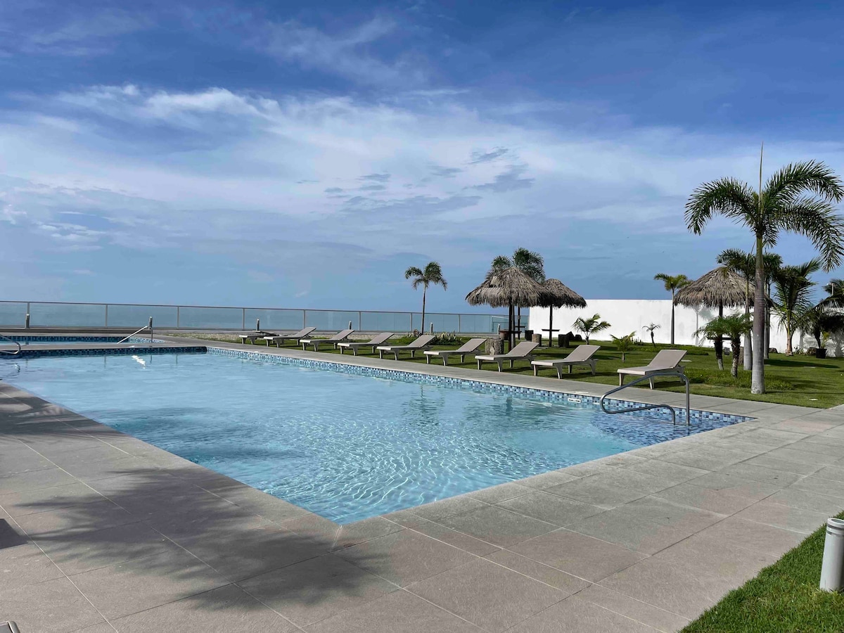 A relaxing pool area features clear blue water with lounging chairs surrounding it. Umbrellas provide shade near the water's edge, and palm trees sway gently in the background. The horizon showcases a serene view of the ocean under a partly cloudy sky.