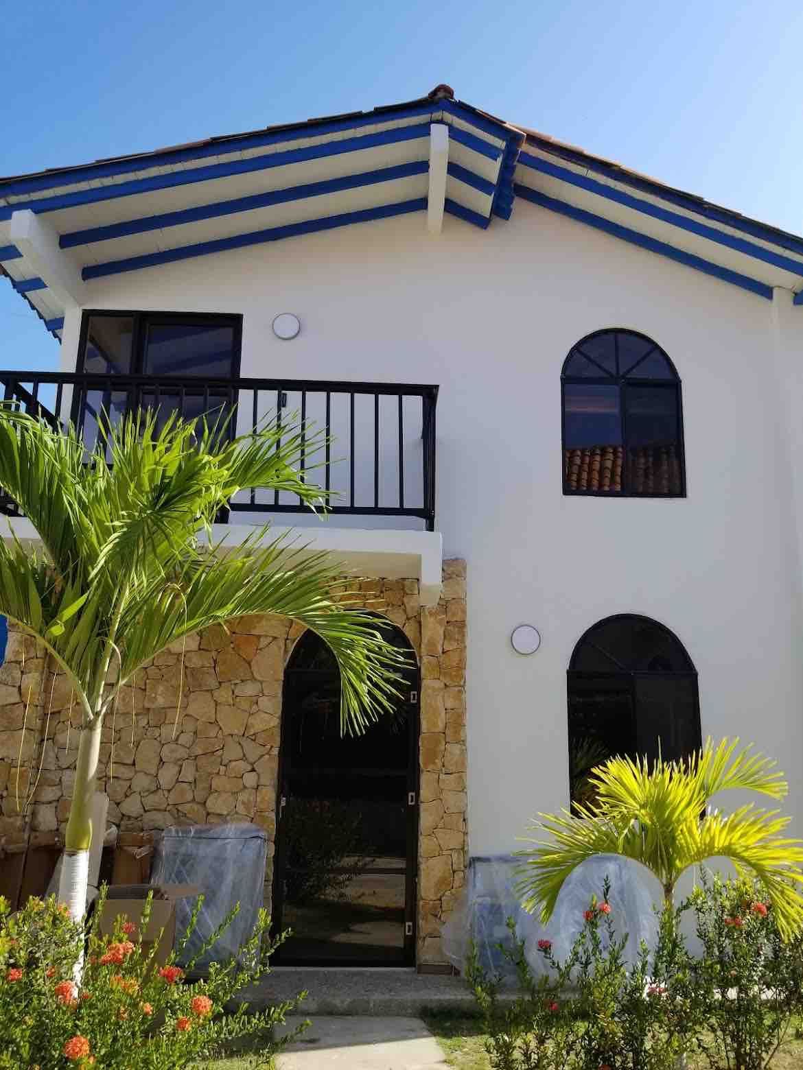 The exterior showcases a modern two-story cabin with an arched entrance and large windows. A balcony extends from the upper level, framed by decorative blue beams. Lush green palm trees and vibrant flowering plants are positioned near the entrance, enhancing the inviting atmosphere.