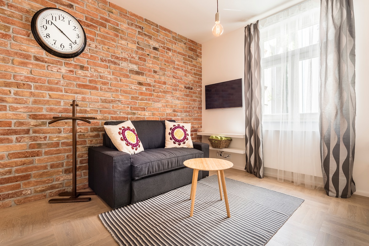 A cozy seating area features a dark sofa adorned with patterned pillows. A round table stands in front on a textured rug, while a wall clock adds character. Natural light filters through sheer curtains, illuminating the exposed brick wall and enhancing the modern design.