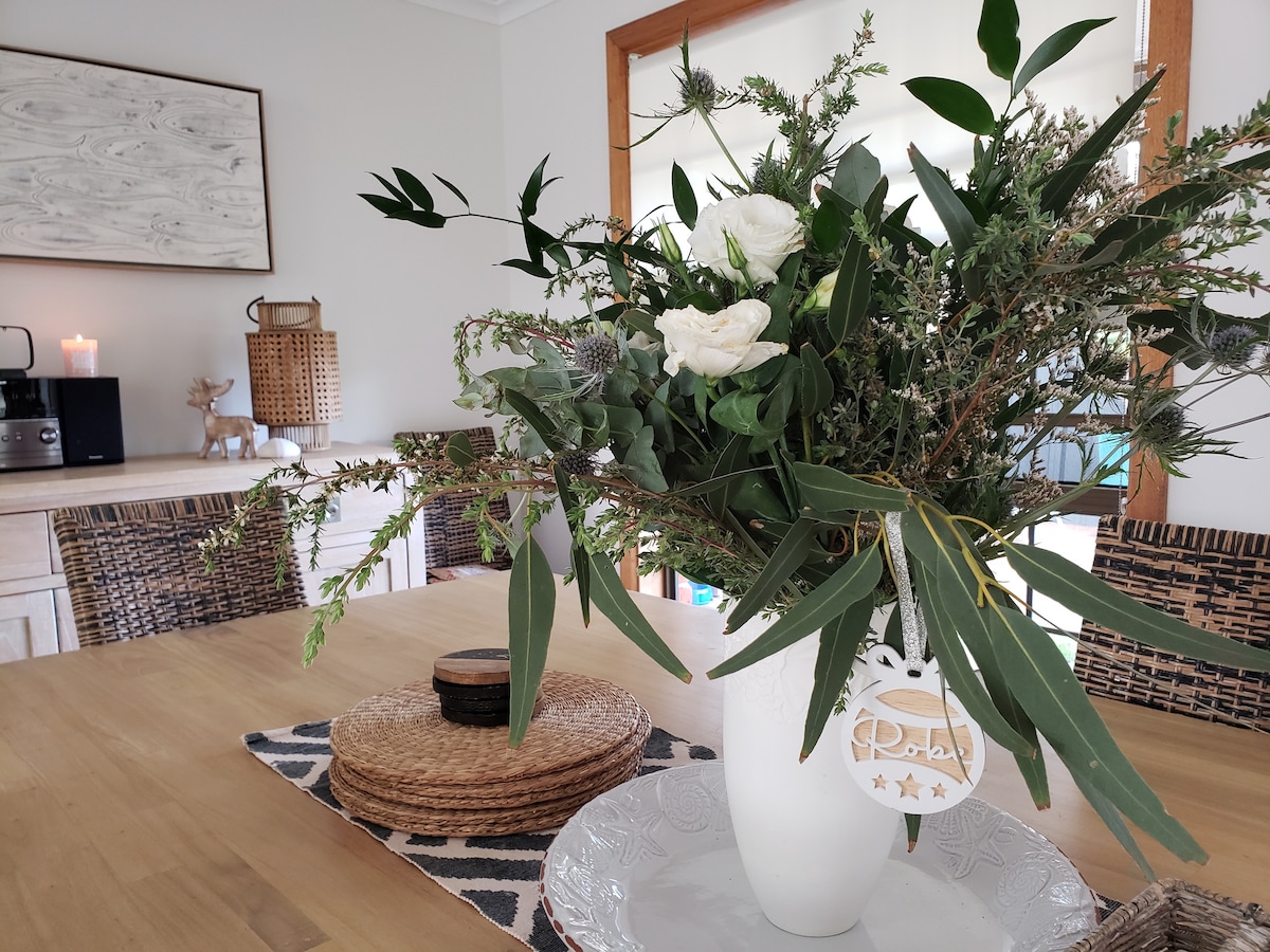 A stylish dining table is adorned with a white vase filled with fresh greenery and flowers. Natural textures are highlighted through wicker placemats and a decorative tray, creating a warm and inviting centerpiece. Soft lighting enhances the overall ambiance of the space.