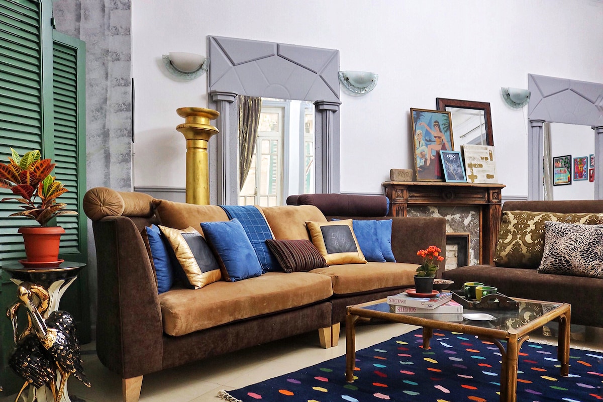A spacious living area is highlighted by a large, comfortable sofa adorned with decorative pillows. A coffee table sits on a patterned rug, with a plant placed atop it. Reflections from mirrors and light coming through windows create a bright and inviting atmosphere.