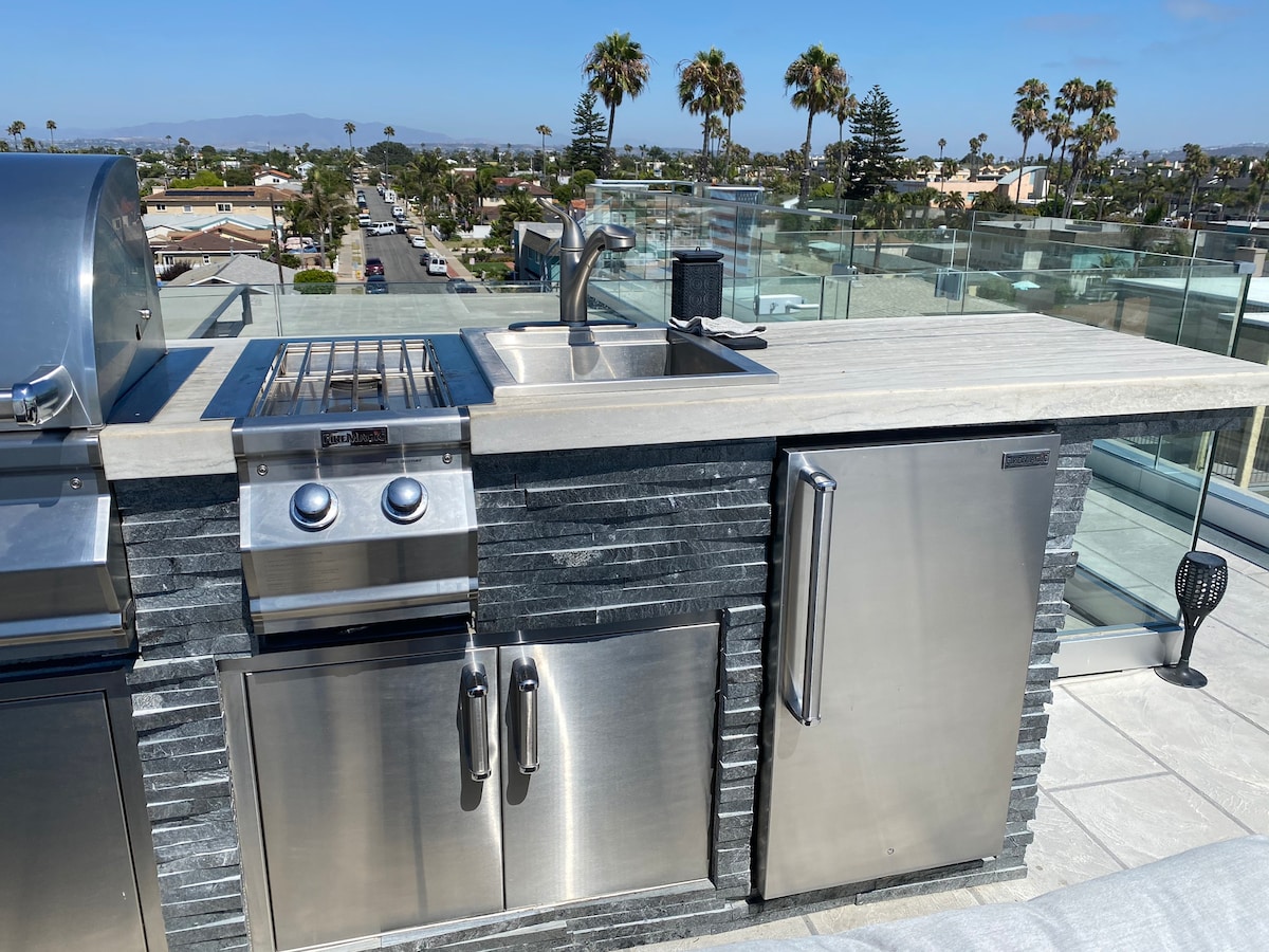 The rooftop deck features a built-in grill, sink, and a refrigerator, all crafted with stainless steel accents. The stone counter provides a modern aesthetic, while a clear view of the coastal landscape is visible in the background, surrounded by palm trees and blue skies.
